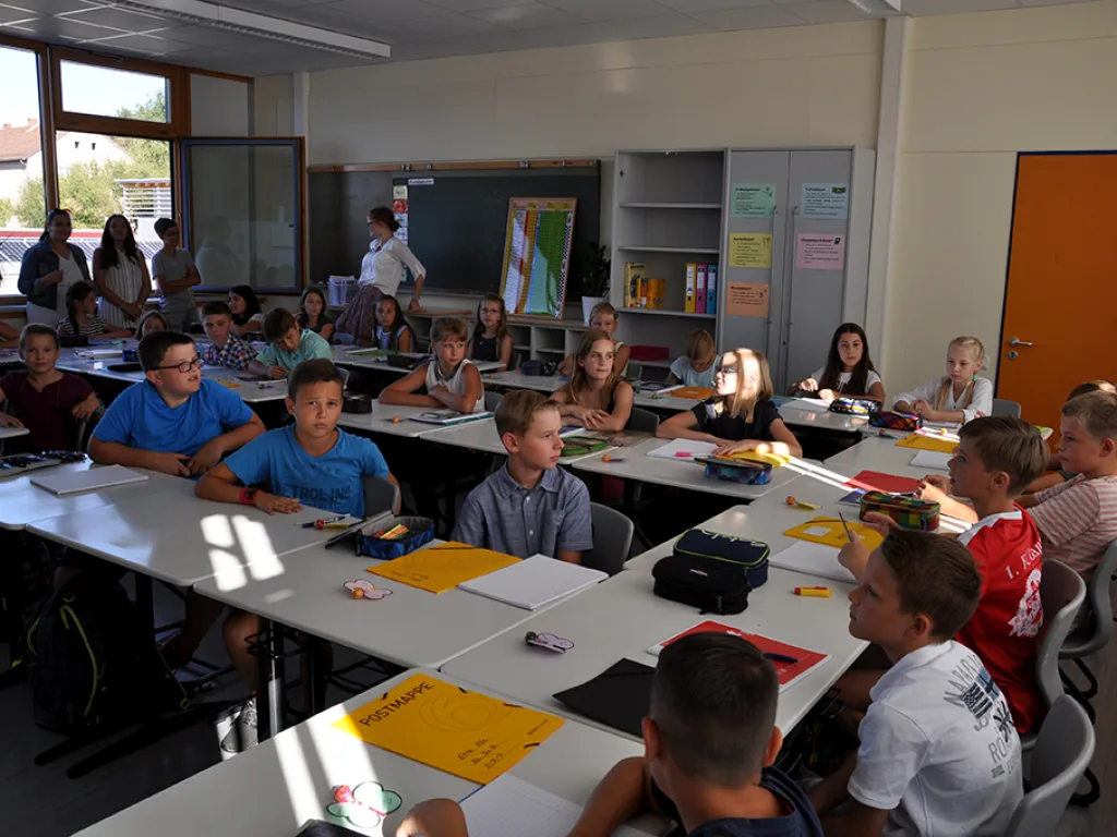 Kinder sitzen im Klassenraum, eine Lehrkraft steht vorn an der Tafel.